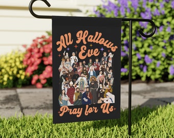 Könnte beinhalten: Schwarze Gartenflagge mit dem Text "All Hallows Eve Pray for Us" in orangefarbener Schrift. Die Flagge zeigt eine farbenfrohe Illustration verschiedener religiöser Figuren. Die Flagge wird auf einem schwarzen Metallständer in einem Garten gezeigt.