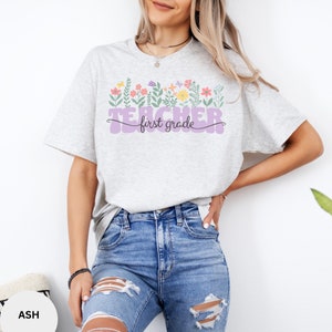 May include: A light grey t-shirt with a purple "Teacher first grade" graphic. The graphic is surrounded by colourful flowers.