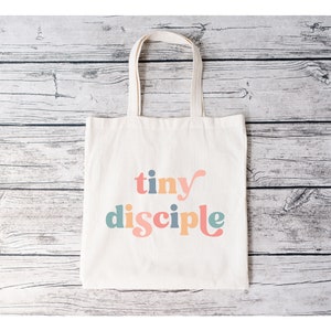 May include: A white canvas tote bag with the text "tiny disciple" in colorful, stylized lettering.