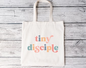 Può includere: Una borsa di tela bianca con il testo "tiny disciple" in lettere colorate e stilizzate.