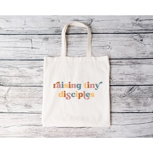 以下が含まれることがあります： カラフルなスタイリッシュな文字で「raising tiny disciples」と書かれた白いキャンバス地のトートバッグです。