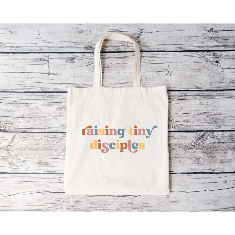 以下が含まれることがあります： カラフルなスタイリッシュな文字で「raising tiny disciples」と書かれた白いキャンバス地のトートバッグです。