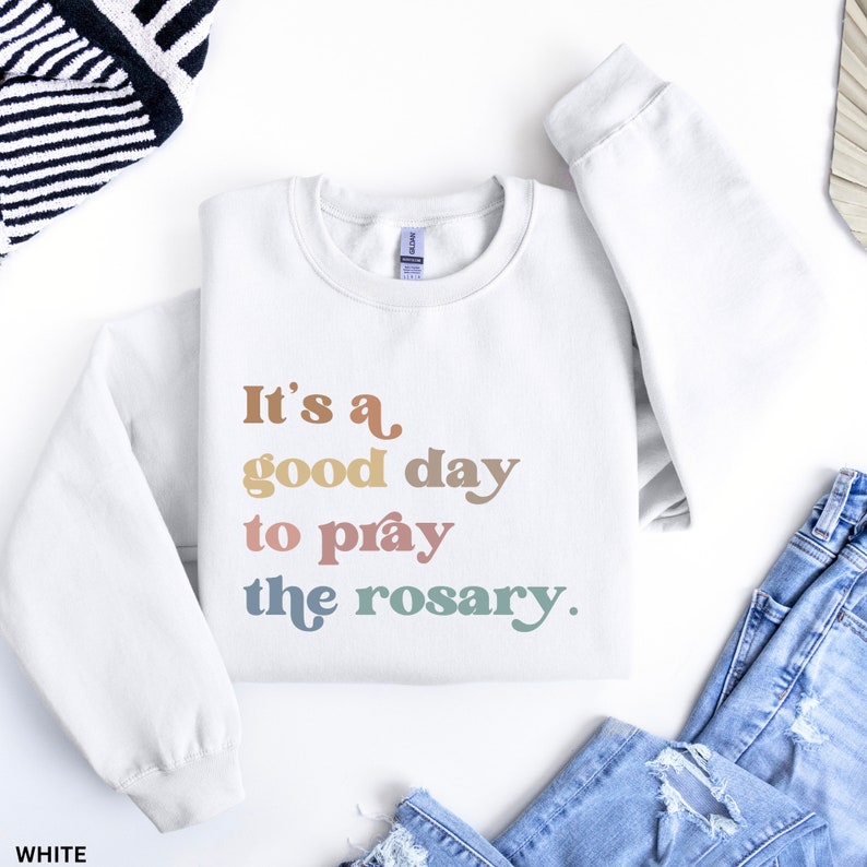 May include: White sweatshirt with the text "It's a good day to pray the rosary." in rainbow colors.
