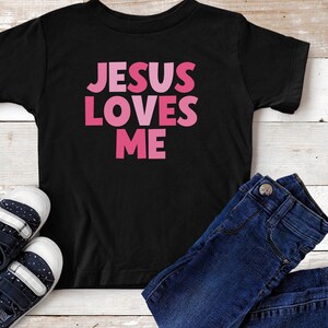 May include: Black t-shirt with the text "JESUS LOVES ME" in pink letters. The letters are in a gradient of pink shades.