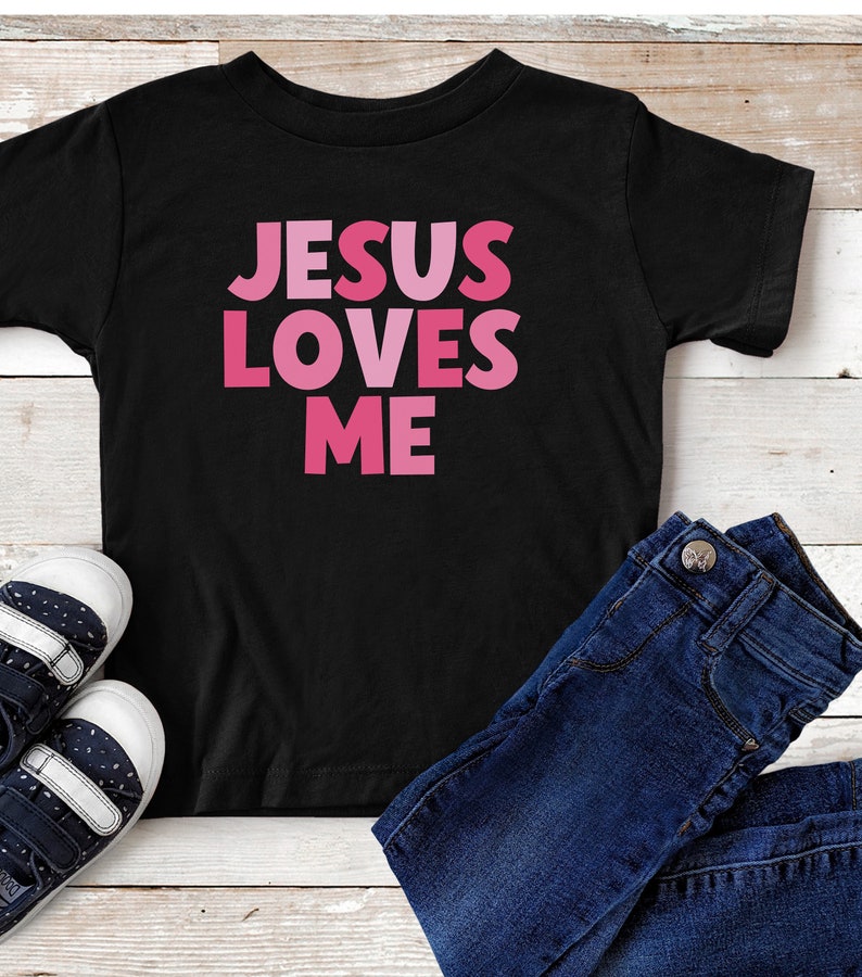 May include: Black t-shirt with the text "JESUS LOVES ME" in pink letters. The letters are in a gradient of pink shades.