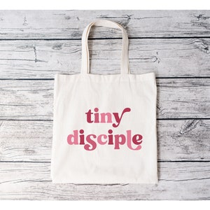 May include: A white canvas tote bag with pink text that reads "tiny disciple".