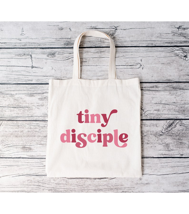 May include: A white canvas tote bag with pink text that reads "tiny disciple".