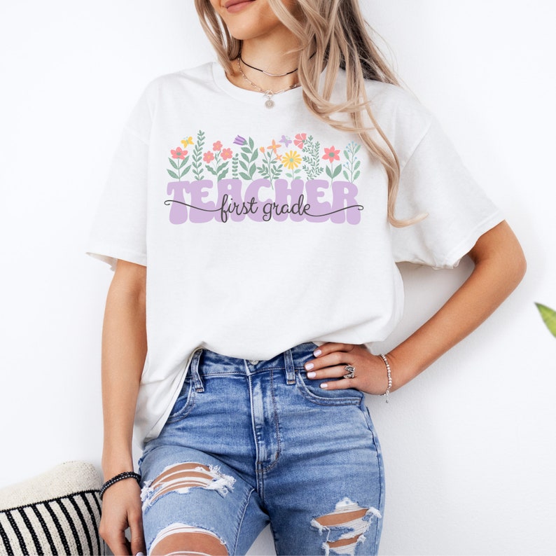 May include: White t-shirt with a purple "Teacher first grade" design and a row of colourful flowers above the text.