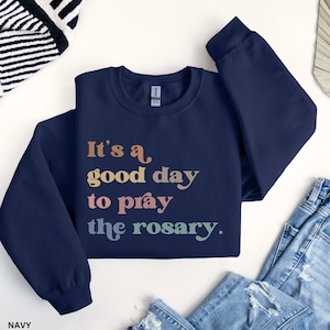 May include: Navy blue sweatshirt with the text "It's a good day to pray the rosary." in rainbow colors.