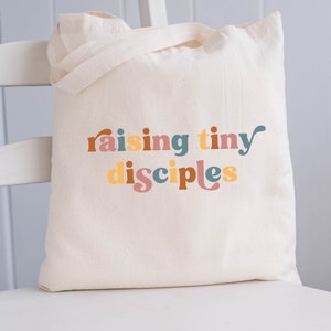 以下が含まれることがあります： レトロなスタイルのカラフルな文字で「raising tiny disciples」と書かれた白いキャンバス地のトートバッグです。