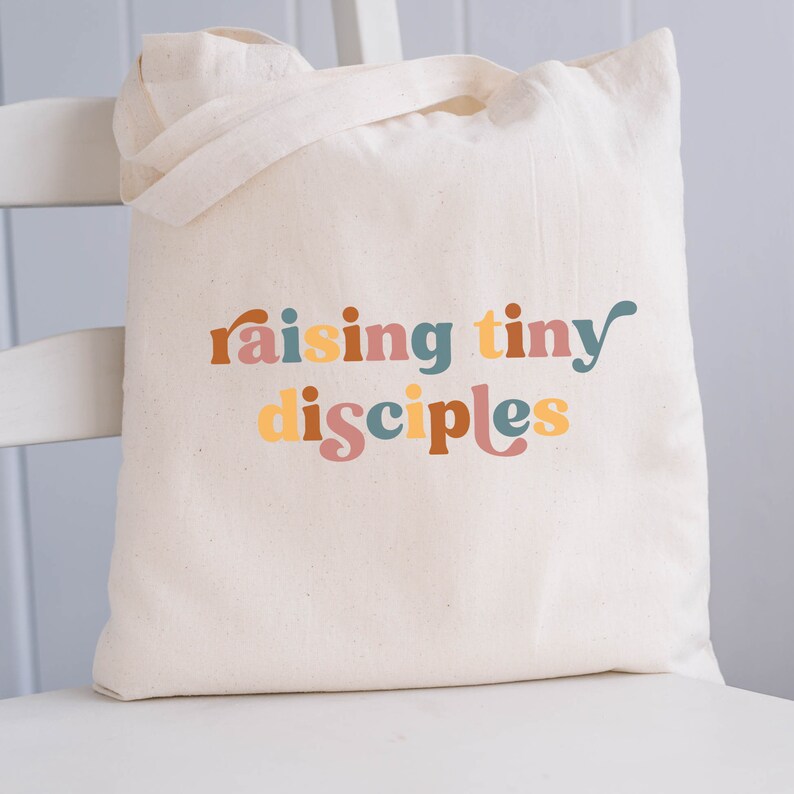 以下が含まれることがあります： レトロなスタイルのカラフルな文字で「raising tiny disciples」と書かれた白いキャンバス地のトートバッグです。