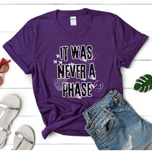 Może przedstawiać: Fioletowy T-shirt z napisem "It Was Never A Phase" w kolorze białym z przetartym wzorem graficznym.