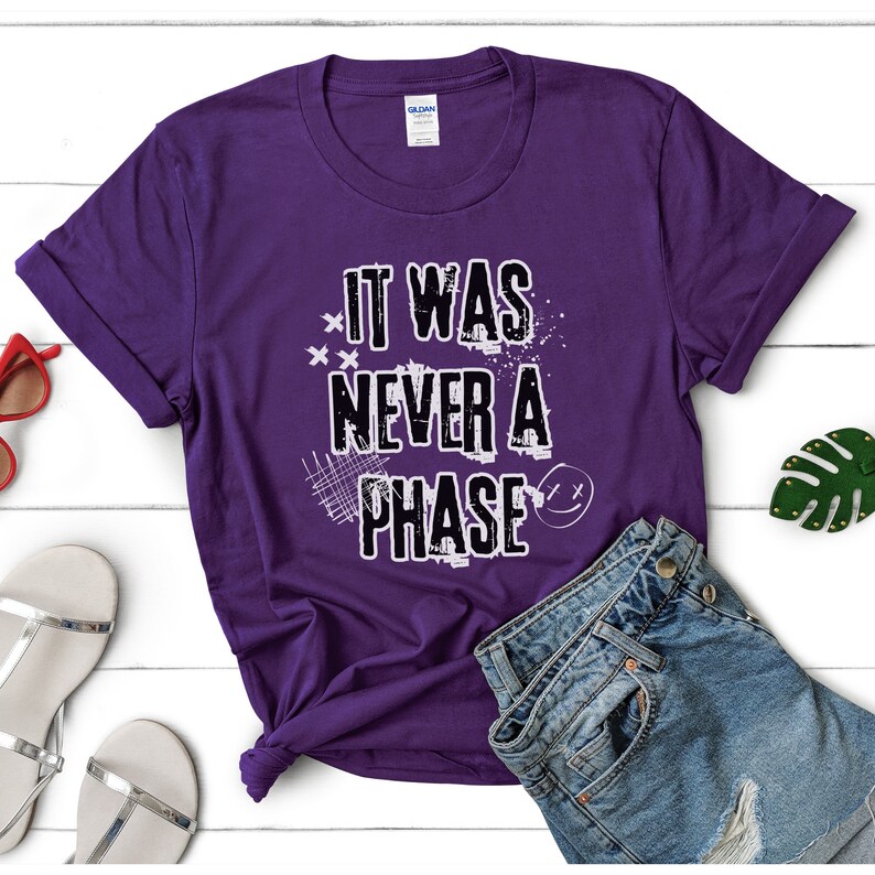Może przedstawiać: Fioletowy T-shirt z napisem "It Was Never A Phase" w kolorze białym z przetartym wzorem graficznym.
