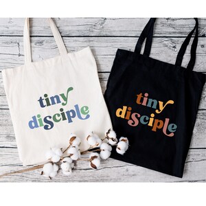 May include: Two tote bags with the text "tiny disciple" printed on them. The bag on the left is white with green, blue, and brown lettering. The bag on the right is black with orange, brown, and green lettering.