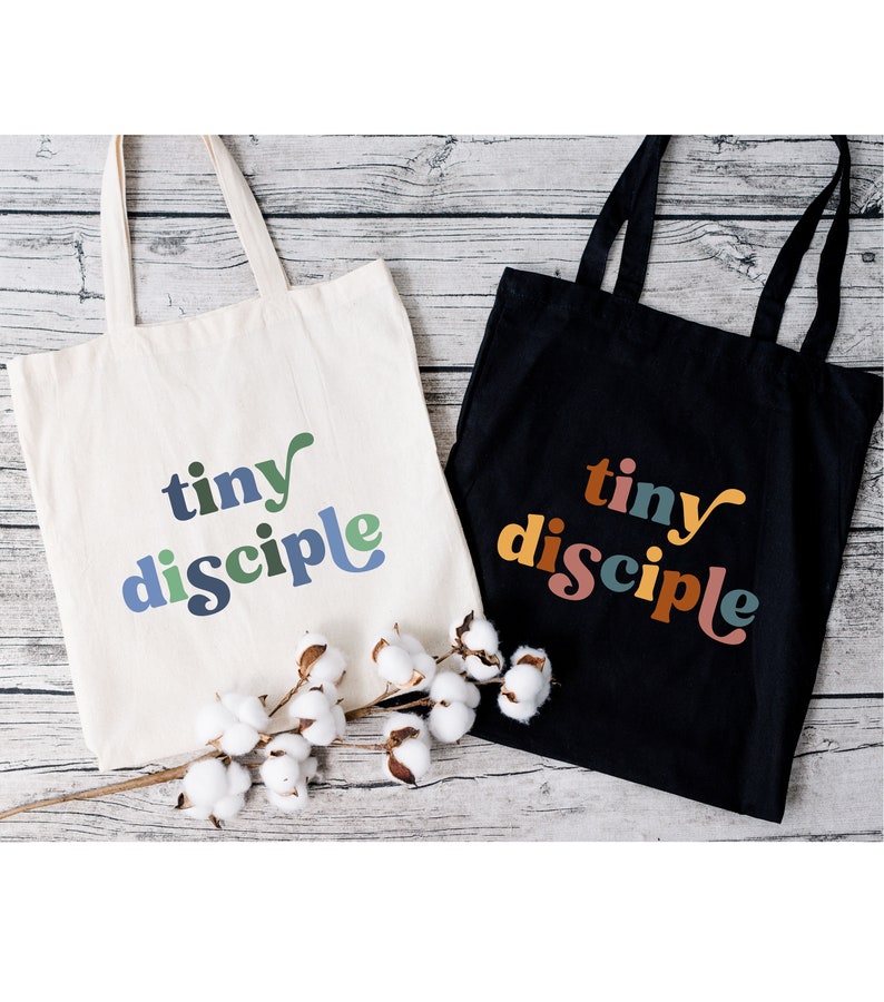 May include: Two tote bags with the text "tiny disciple" printed on them. The bag on the left is white with green, blue, and brown lettering. The bag on the right is black with orange, brown, and green lettering.