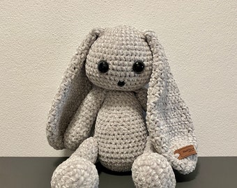 May include: A handmade, gray crocheted bunny with long, floppy ears and large black eyes. The plush toy has a soft, textured appearance and is sitting upright. A small brown tag is attached to the bunny's foot.