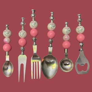 Puede incluir: Conjunto de seis utensilios de plata con cuentas de corazón rosa y blanco. Los utensilios incluyen una cuchara, un tenedor, una cuchara de servir, un tenedor de servir y un abridor de botellas.