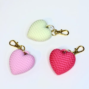 May include: Three heart-shaped keychains in pastel colors: light green, pink, and red. Each keychain has a textured surface and a gold-colored clasp. The keychains are arranged on a white background.