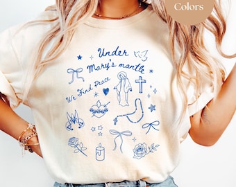 May include: A white t-shirt with a blue design that says "Under Mary's mantle We Find Peace" and includes a variety of religious symbols. The shirt is shown on a model wearing blue jeans. The color of the shirt is ivory.