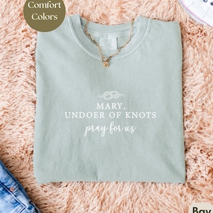 May include: A light green Comfort Colors t-shirt with the text "MARY, UNDOER OF KNOTS pray for us" in white. A gold necklace with a pendant is on the shirt. The shirt is on a pink fluffy surface.