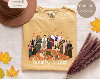 May include: A mustard yellow t-shirt with a graphic of a group of saints surrounded by autumn leaves. The text "Saintly Season" is printed below the graphic.