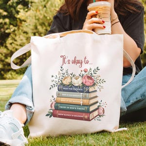 May include: Cream-colored tote bag with the text "It's okay to..." and a stack of books with floral accents. The books have titles like "Pray for Patience" and "Trust in God's Timing". The bag has white handles.