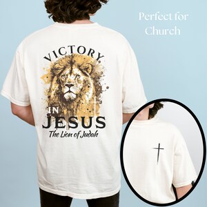May include: A white t-shirt with a lion graphic and the text "Victory in Jesus The Lion of Judah". The back of the shirt has a small cross graphic.