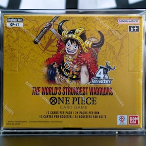 May include: A sealed One Piece Card Game booster box, The World's Strongest Warriors, encased in a clear acrylic display. The yellow box features a character with horns, the 4th Anniversary logo, and text indicating 12 cards per pack, 24 packs per box.