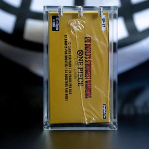 May include: A sealed One Piece Card Game booster box, labeled The World's Strongest Warriors, encased in a clear acrylic display case. The gold box features black text indicating 131+4 types, 12 cards per pack, 24 packs per box, and English Ver.