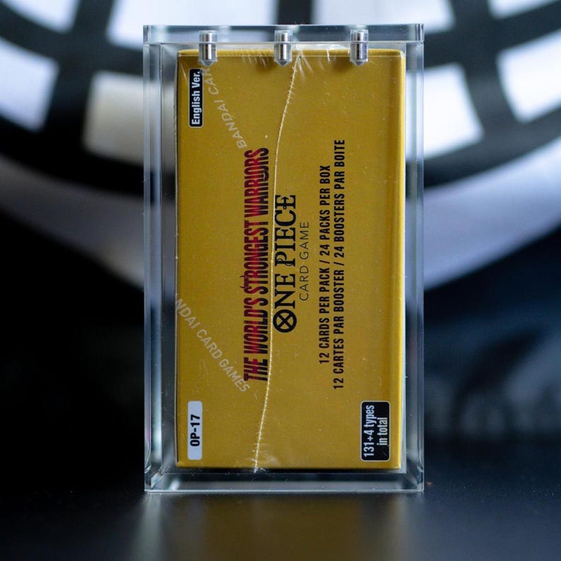 May include: A sealed gold One Piece Card Game booster box, labeled The World's Strongest Warriors, encased in a clear acrylic display case. The box features text indicating 12 cards per pack, 24 packs per box, and 131 plus 4 types in total.