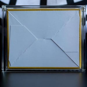 May include: A rectangular object encased in a clear acrylic frame. The object features a white paper envelope flap centered on a solid mustard yellow background. The entire assembly is sealed in a thin, transparent plastic film wrap.