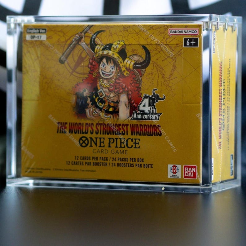 May include: A One Piece Card Game booster box titled The World's Strongest Warriors, 4th Anniversary edition, encased in a clear acrylic display case. The yellow box features an illustration of a character in a horned helmet and text in English.