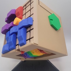 May include: A beige cube puzzle with a green hexagonal piece on one side. The cube has a grid pattern on one side and a circular pattern with numbers on another side. The cube is on a black stand with the word "nostalgia" printed on it.