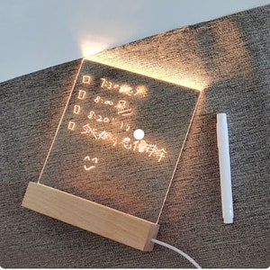 K&ouml;nnte beinhalten: Ein leuchtendes Acryl-Message-Board mit einem Holzsockel. Das Board hat einen wei&szlig;en Marker und handschriftlichen Text, der "7:30 Aufwachen", "8:00", "8:20", "Fr&uuml;hst&uuml;ck" und "18:00 Uhr" lautet.