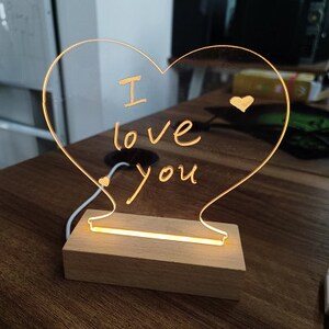 K&ouml;nnte beinhalten: Ein herzf&ouml;rmiges Acrylschild mit Holzsockel und warmwei&szlig;em LED-Licht. Das Schild tr&auml;gt die Aufschrift "Ich liebe dich" in handschriftlicher Schrift.