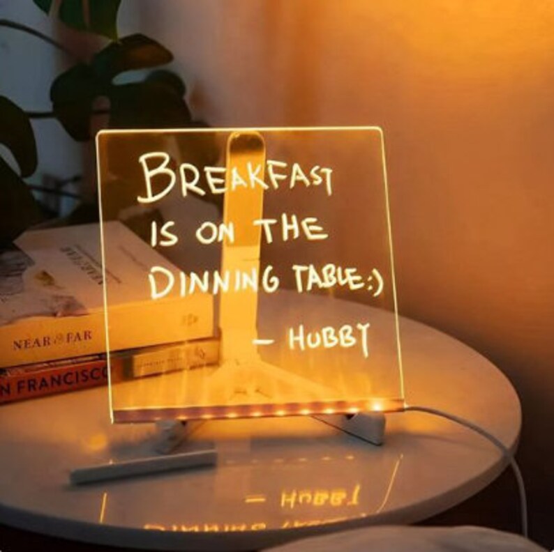 K&ouml;nnte beinhalten: Ein klares Acrylschild mit einem warmen wei&szlig;en LED-Lichtrahmen. Das Schild tr&auml;gt eine handgeschriebene Nachricht mit dem Text "Breakfast is on the dining table:) - Hubby".