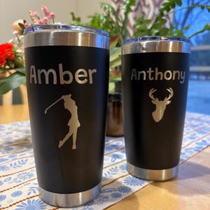 May include: Two black stainless steel tumblers with personalized names, "Amber" and "Anthony". The tumbler on the left has a golfer silhouette, and the tumbler on the right has a deer silhouette.