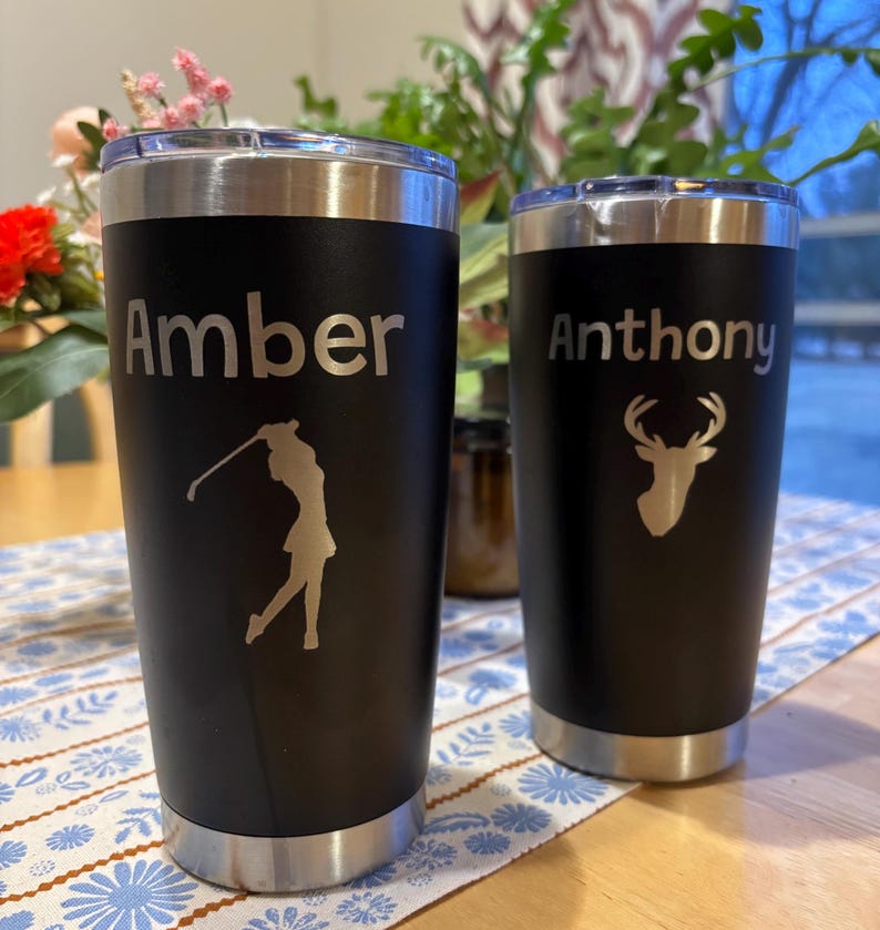 May include: Two black stainless steel tumblers with personalized names, "Amber" and "Anthony". The tumbler on the left has a golfer silhouette, and the tumbler on the right has a deer silhouette.