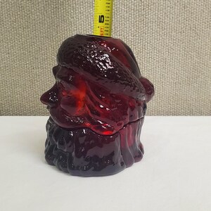 May include: A deep red glass Santa Claus head container with a textured hat and beard. A yellow measuring tape is positioned behind the glass, showing the object stands approximately 5 inches tall from the base to the top opening of the glass.