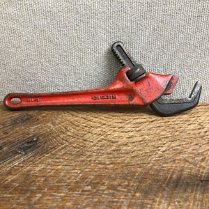 May include: A vintage red Ridgid pipe wrench, a hand tool for gripping and turning pipes. The wrench is made of metal and has a black jaw. The tool is lying on a wooden surface.