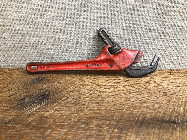May include: A vintage red Ridgid pipe wrench, a hand tool for gripping and turning pipes. The wrench is made of metal and has a black jaw. The tool is lying on a wooden surface.