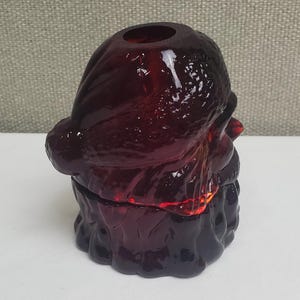 May include: A deep red glass figurine shaped like a Santa Claus head, featuring a textured hat and beard with a hollow opening at the top. The translucent material catches light, highlighting the molded facial details and the rounded form of the figure.