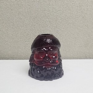 May include: A deep red translucent glass votive candle holder shaped like the head of Santa Claus, featuring a textured hat, a detailed face with a mustache and beard, and a circular opening at the top for a candle or small decorative item.