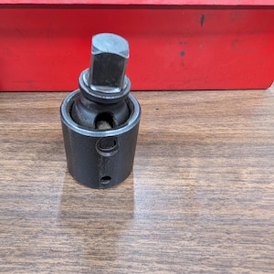 May include: A black socket wrench adapter with a square drive end and a cylindrical body. The adapter is made of metal and has two small holes on the side. A red toolbox is in the background.