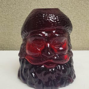 May include: A deep red glass vessel shaped like the head of Santa Claus, featuring a textured hat and a detailed, flowing beard. The translucent material catches the light, highlighting the molded facial features and the open top design.