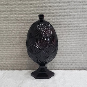 May include: A black glass candy dish with a lid. The dish is decorated with a cut glass pattern of leaves and has a pedestal base.
