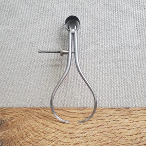 May include: A vintage silver-colored caliper tool with curved legs and a screw adjustment. The tool has a circular top and is set against a textured gray background and a wooden surface.