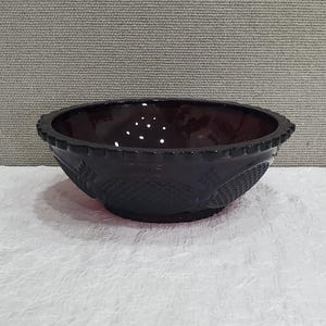 May include: A dark purple glass bowl with a decorative rim and patterned sides. The bowl has a diamond and floral design. The bowl is sitting on a white surface.