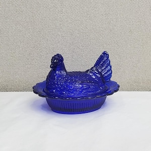 May include: A cobalt blue hen-shaped covered dish. The dish features a detailed hen design on the lid and a scalloped edge. The dish is made of glass and is sitting on a white surface.