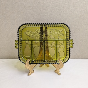 May include: A rectangular, olive-green glass serving dish with four compartments and a decorative fruit design. The dish has a scalloped edge and small handles. It is displayed on a gold-colored metal stand.